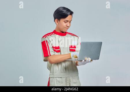 Portrait of smiling handsome mechanic man wearing uniform holding and using laptop on gray background Stock Photo