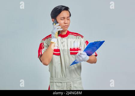 Portrait of young Asian mechanic talking on phone and holding clipboard with confused expression isolated on gray background Stock Photo