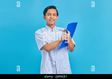 Smiling young male nurse taking notes on clipboard and looking at camera on blue background Stock Photo