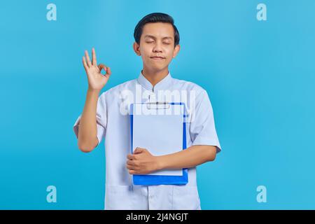 Smiling young male nurse showing clipboard and making ok gesture with closed eyes on blue background Stock Photo