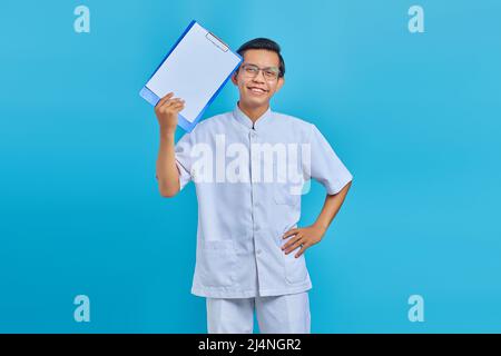 Cheerful young male nurse showing clipboard and looking at camera on blue background Stock Photo