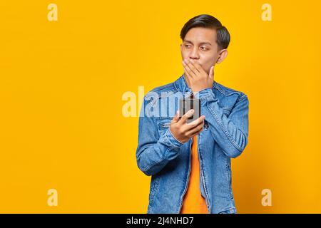 Shocked asian man covering mouth with hands and holding smartphone over yellow background Stock Photo