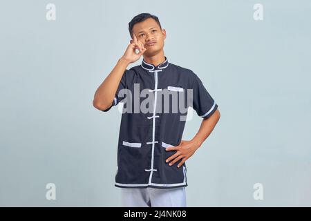 Portrait of pensive Asian young man wearing taekwondo kimono thinking about questions with hands on head isolated on gray background Stock Photo