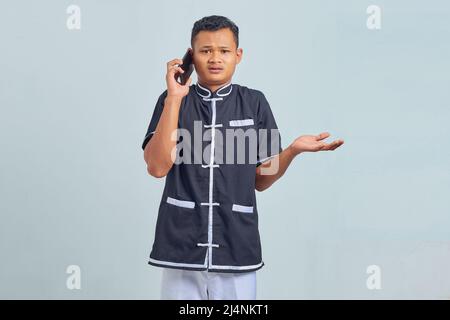 Portrait of confused asian young man wearing karate uniform talking on smartphone on gray background Stock Photo