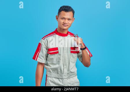 Image of smiling Asian handsome man wearing mechanical uniform holding wrench isolated on blue background Stock Photo