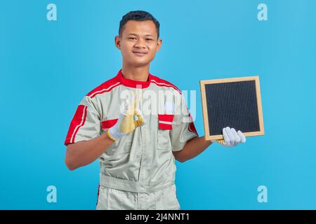 Portrait of cheerful young Asian mechanic holding blank board and doing ok gesture over blue background Stock Photo