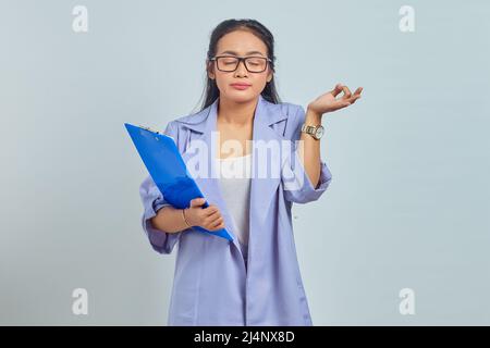 Portrait of beautiful young Asian woman holding document folder and standing in lotus pose, trying to meditate yoga, close eyes isolated on purple bac Stock Photo