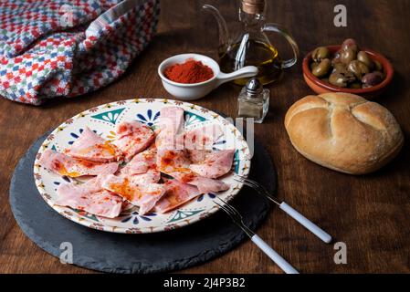 Plate with pieces of roasted ham, with oil, pepper and sweet paprika, accompanied by its ingredients and assorted bread. On a wooden background. Stock Photo