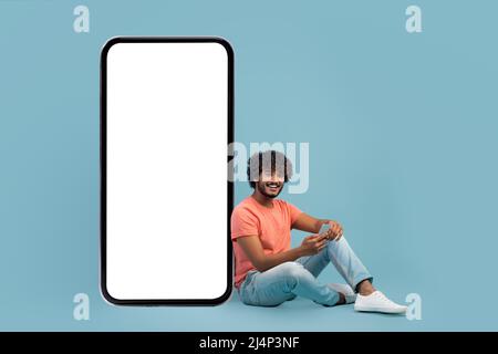 Handsome indian guy sitting by huge smartphone, using cell phone Stock Photo