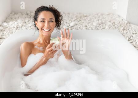 Attractive young woman lying in hot bubble bath, applying cream or hair mask at home, free space Stock Photo