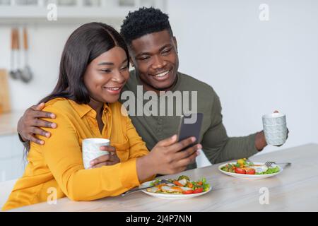 Young Black Man And Woman Using Smartphone While Enjoying Breakfast In Kitchen Stock Photo