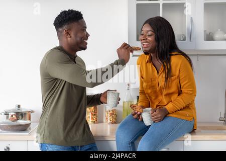 Cheerful Young Black Couple Eating Snacks In Cozy Kitchen Together Stock Photo