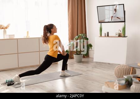 Female Doing Forward Lunge Stretch Watching Workout On Television Indoors Stock Photo