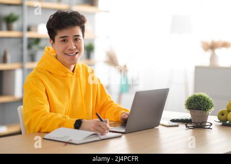 Smiling asian man working on laptop and writing Stock Photo
