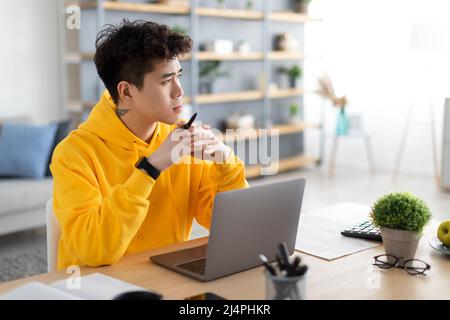 Pensive Asian man using laptop sitting at desk in office Stock Photo