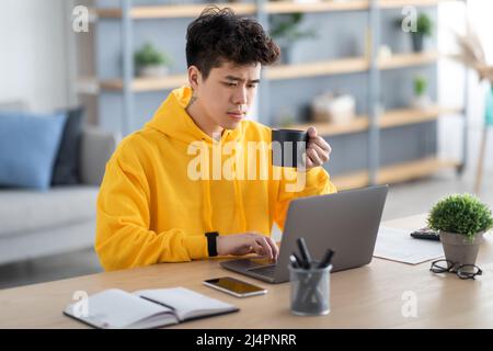 Serious asian man watching video on pc, drinking hot coffee Stock Photo