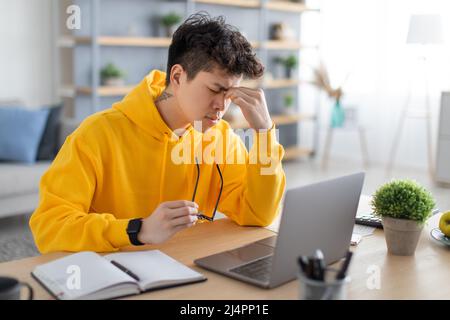 Tired Asian guy sitting at desk with pc, rubbing nosebridge Stock Photo