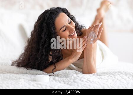 Satisfied young caucasian brunette lady with long curly hair in towel applies cream on hand, lies on bed Stock Photo