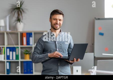 Portrait of handsome mature male office worker with laptop in hands standing in office and smiling at camera Stock Photo