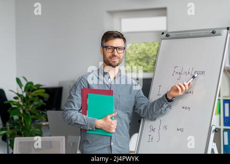 Distance learning concept. Mature male tutor giving English class, writing on blackboard in office interior Stock Photo