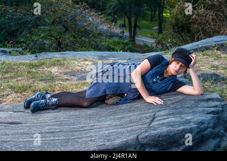 Dressing in a blue sleeveless long blouse, black leggings and boots, wearing a black corduroy cop, a young Chinese girl is lying on rocks, one hand su Stock Photo