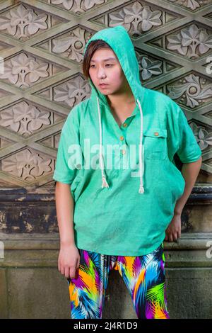 Dressing in a green pull over hoodie,  colorful pants, a young Chinese girl is standing against an old fashion style wall, lost in thought. Stock Photo