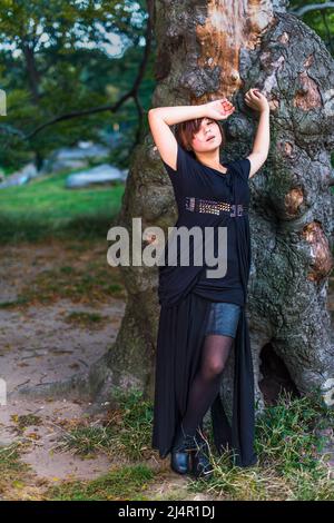 A Chinese girl, dressing in dark, is tired, unhappy, standing by an old tree trunk in the evening. Stock Photo