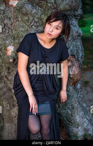 A Chinese girl, dressing in dark, is leaning against an old tree trunk,  tired and relaxing Stock Photo