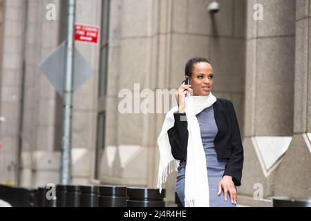 Dressing in a woolen cropped jacket, a fitted dress and a long scarf, a young black woman is sitting outside and smilingly listening a phone call. Stock Photo