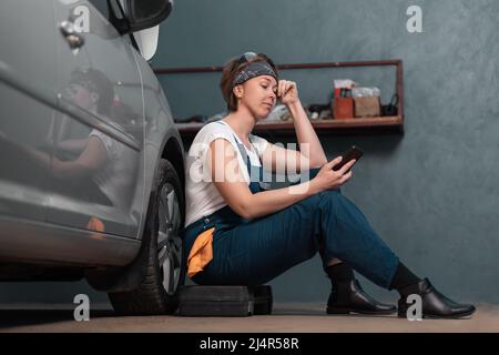 Break time at job. Caucasian young woman mechanic is sitting leaning on a car in an auto repair shop and using cellphone. The concept of women's work Stock Photo