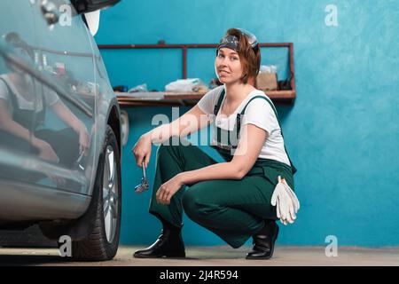 Young smiling female mechanic in blue coveralls squatting near tire and holding ratchet wrench. The concept of women's work in men's professions and e Stock Photo