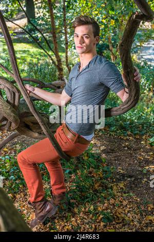 Dressing in a gray long sleeves with roll-tab Henley shirt,  red jeans and brown leather boot shoes, a young guy is sitting on a rattan, relaxing outs Stock Photo