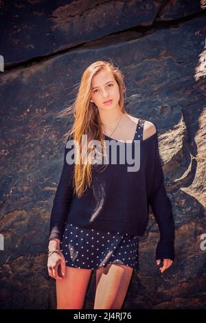 American Teenage Girl with long blonde hair, wearing black knit sweater, patterned skirt, standing against rocky wall at Central Park, New York, charm Stock Photo