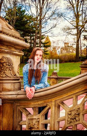American teenage girl wearing Denim jacket, bending over, arms on old fashion style fence at Central Park, New York, smiling, looking at you. Instagra Stock Photo