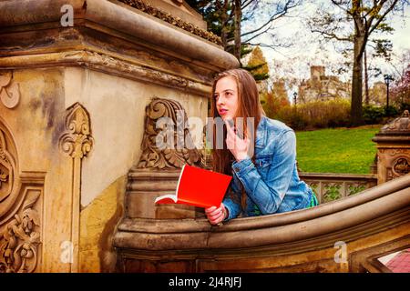 American teenage girl wearing Denim jacket, reading ed book outside on campus in New York, thinking. Instagram Filtered Look Stock Photo