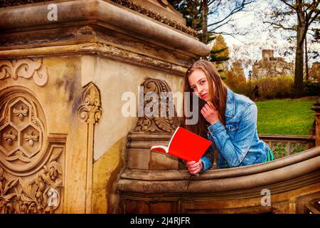 American teenage girl wearing Denim jacket, reading ed book outside on campus in New York, thinking. Instagram Filtered Look Stock Photo
