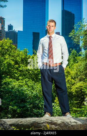 Young American Businessman wearing white long sleeve shirt, black pants, pattern tie, two hands in pockets, standing in front of business district in Stock Photo