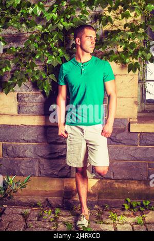 Young man waiting. Wearing a green short sleeve Henley shirt, light yellow shorts, leather casual shoes, bending a leg, a young guy is standing agains Stock Photo