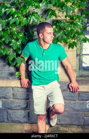 Young man waiting. Wearing a green short sleeve Henley shirt, light yellow shorts, bending a leg, a young guy is standing against the wall with green Stock Photo