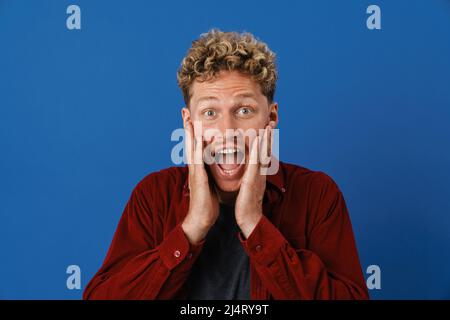 Young blonde man with curly hair expressing surprise at camera isolated over blue wall Stock Photo