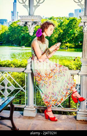 Texting Anywhere. Dressing in sleeveless, long dress, red sandals shoes, a teenage girl is sitting inside a pavilion, hands holding a smart phone, typ Stock Photo