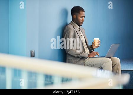 Vibrant side view portrait of black young businessman using laptop while working in blue office space Stock Photo