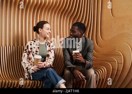 Portrait of to young coworkers chatting while relaxing at lounge in modern office space with wooden panelling Stock Photo