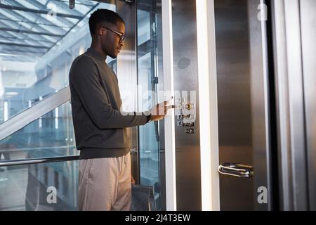 Graphic side view at young black man pushing button in glass elevator in modern office building, copy space Stock Photo