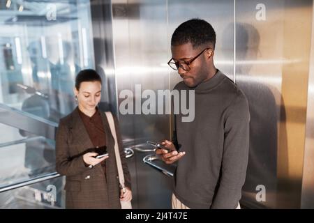 Side view portrait of young black businessman using smartphone in elevator at modern office building with coworker Stock Photo