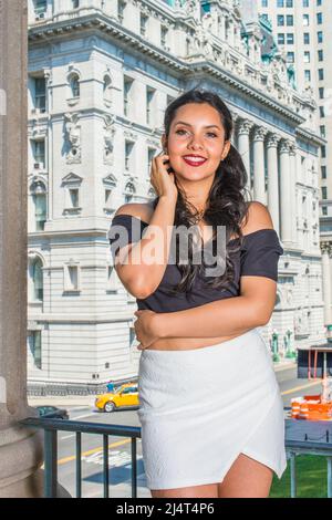 Portrait of College Student. Dressing in black, short sleeve top, white short wrap skirt, a young pretty lady with long curly hair is standing in the Stock Photo