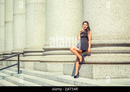 College Student. Dressing in black, sleeveless top, short pants, high heels, a young pretty lady with long curly hair is standing against columns outs Stock Photo