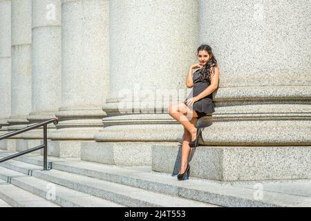College Student. Dressing in black, sleeveless top, short pants, high heels, a young pretty lady with long curly hair is standing against columns outs Stock Photo