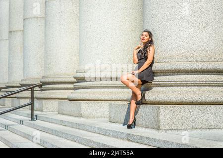 College Student. Dressing in black, sleeveless top, short pants, high heels, a young pretty lady with long curly hair is standing against a columns ou Stock Photo