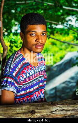 Portrait of Young Black Man. Wearing a colorful pattern shirt, necklace, a young handsome guy is standing by a wooden fence, charmingly looking at you Stock Photo